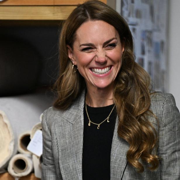 Prinzessin Kate mit langen, gewellten Haaren lacht herzlich und trägt einen karierten Blazer sowie eine goldene Kette mit Anhängern.