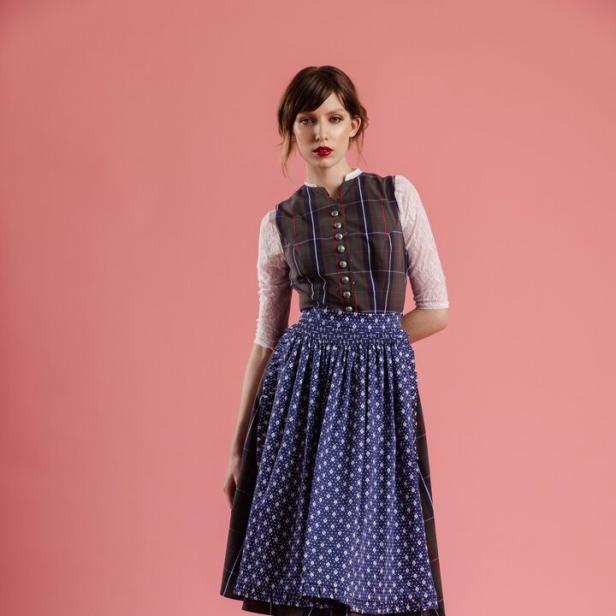 Eine Frau in einem Dirndl mit blauem Rock und karierter Weste vor einem rosa Hintergrund.