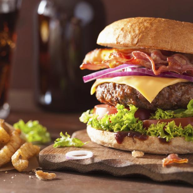 Ein Hamburger mit Speck, Käse, Tomaten und Salat, angerichtet auf einem Brett, daneben Pommes Frittes.