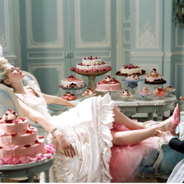 Eine Frau im Rokoko-Kleid ruht sich aus, während ein Dienstmädchen ihr die Schuhe anzieht, umgeben von Kuchen.