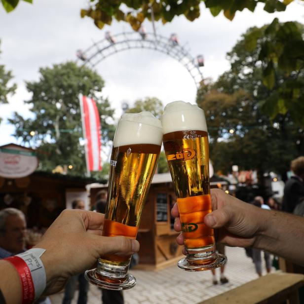 Zwei Hände mit Biergläsern, die gerade zusammenstoßen, im Hintergrund sieht man das Wiener Riesenrad.