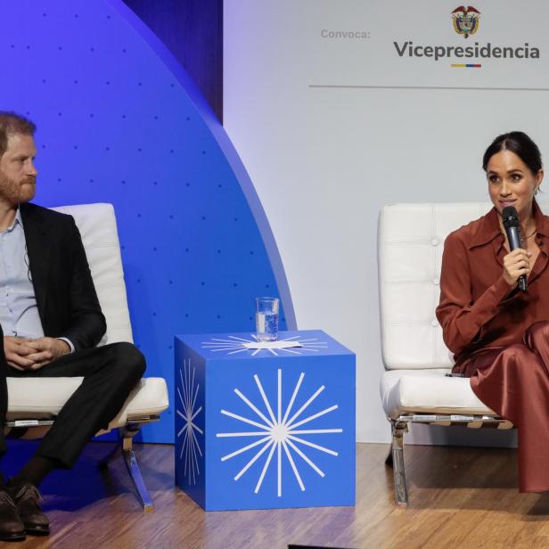 Meghan spricht am Mikrofon, während Harry daneben auf einem weißen Sessel sitzt.