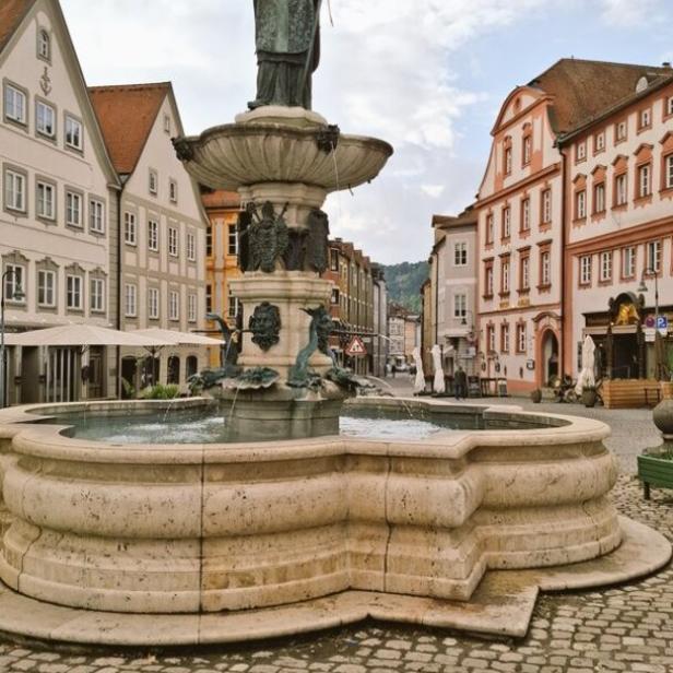 Ein Brunnen mit einer Bischofsstatue auf einem gepflasterten Platz in einer Stadt.