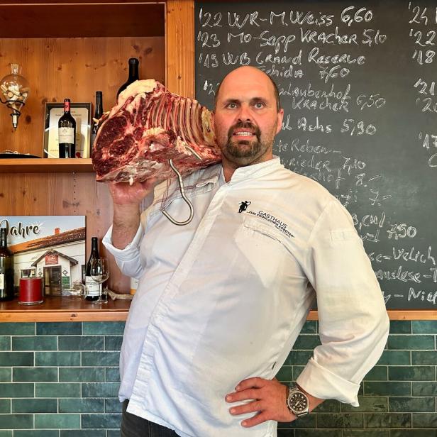 Der Wirt Hannes Tschida trägt ein großes Stück Fleisch auf der Schulter. Auf der Tafel hinter ihm ist die Weinkarte mit Kreide aufgemalt.  