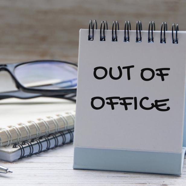 Ein Tischkalender zeigt die Aufschrift „OUT OF OFFICE“, daneben liegen eine Brille, Notizbücher und ein Stift.