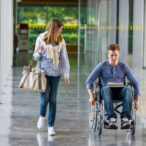 Eine junge Frau geht neben einem jungen Mann im Rollstuhl einen Flur entlang.