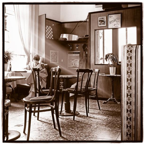 Eine Frau sitzt im Kaffeehaus am Fenster, Sonnenlicht fällt rein. Sie liest Zeitung.