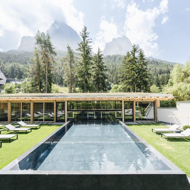 Ein rechteckiger Pool mit Liegestühlen auf einer Rasenfläche vor einer bewaldeten Bergkulisse.