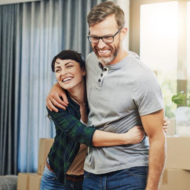 Ein Mann und eine Frau befinden sich in einer Wohnung mit Umzugskartons im Hintergrund und umarmen sich lächelnd.