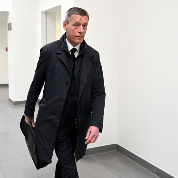 Ein Mann in einem dunklen Mantel geht mit einer Aktentasche einen Flur entlang.