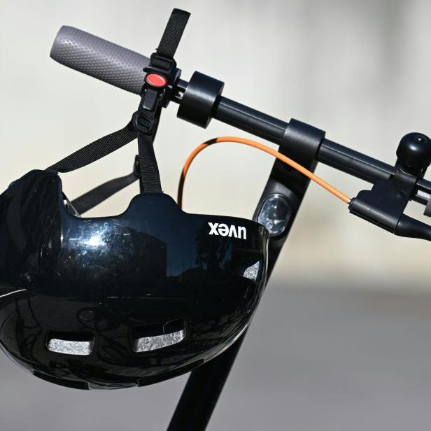 Ein schwarzer Fahrradhelm hängt am Lenker eines E-Scooters mit grauen Griffen und orangefarbenem Kabel.
