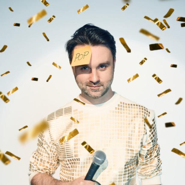 Ein Mann - Dr. Pop - mit Mikrofon und einem Zettel mit der Aufschrift „POP“ auf der Stirn steht im goldenen Konfettiregen.