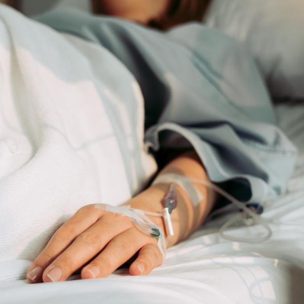 Eine Person liegt im Krankenhausbett, ein Infusionsschlauch ist an ihrem Arm befestigt.