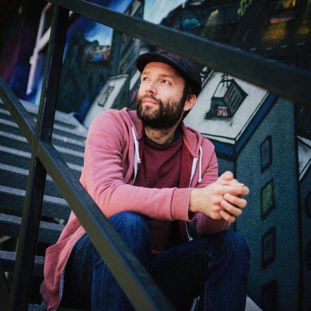 Ein Mann mit Bart und Schirmmütze - Marc-Uwe Kling - sitzt auf einer Treppe vor einer bemalten Wand.