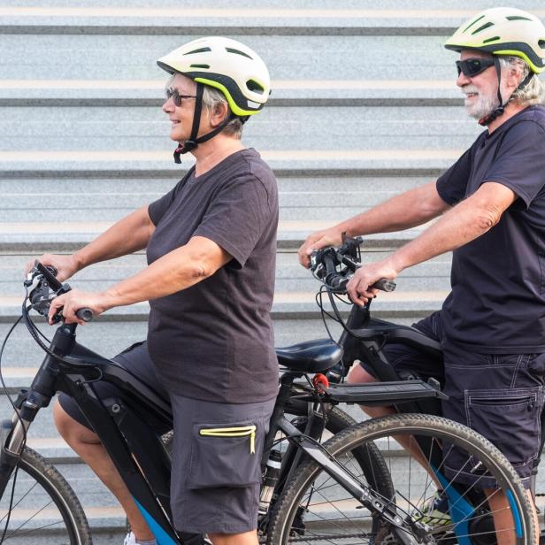 Happy senior couple doing together outdoor activity with electric bicycle. Healthy lifestyle. Yellow helmet, black clothes. Outdoor against a gray metal panel