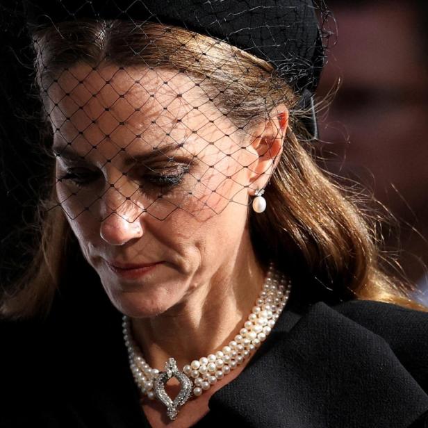 Prinzessin Kate, mit einem Schleierhut und einer Perlenkette, blickt gesenkt.