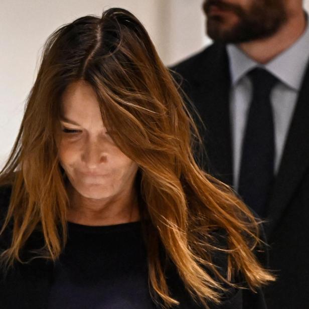 Carla Bruni blickt nach unten, im Hintergrund steht ein Mann im Anzug.