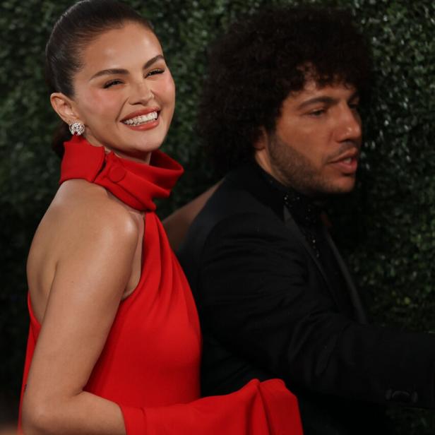 Selena Gomez lächelt in einem roten Kleid steht neben Benny Blanco im Anzug.