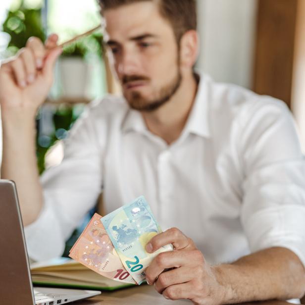 Mann mit Geld in der Hand sitzt vor einem Laptop