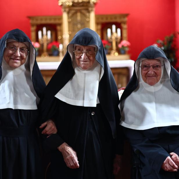 Drei Nonnen in Tracht stehen vor einem roten Altarraum.