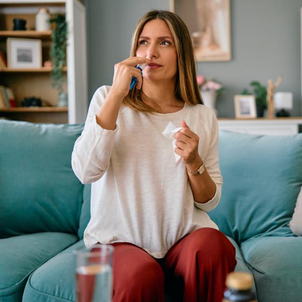 Eine Frau sitzt auf einem Sofa und benutzt Nasenspray, während sie ein Taschentuch in der Hand hält.