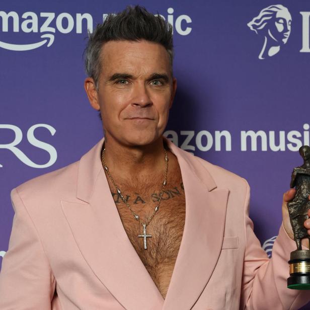 Robbie Williams im rosa Sakko hält eine Auszeichnung vor einem lila Hintergrund mit Amazon Music Logos.