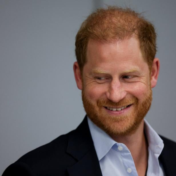 Prinz Harry mit rötlichem Bart und kurzem Haar lächelt und trägt ein dunkles Sakko über einem hellblauen Hemd.