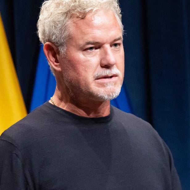 Eric Dane mit grauem Haar und Bart steht vor Flaggen und spricht in ein Mikrofon.