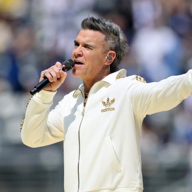 Robbie Williams singt mit Mikrofon in weißer Adidas-Jacke vor verschwommenem Hintergrund.