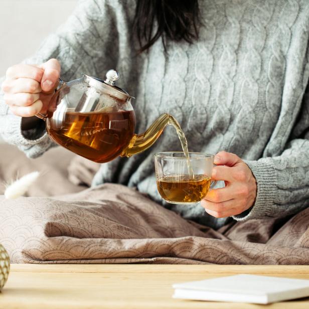 Eine Person gießt Tee aus einer Glaskanne in eine Tasse auf einem Tablett im Bett.