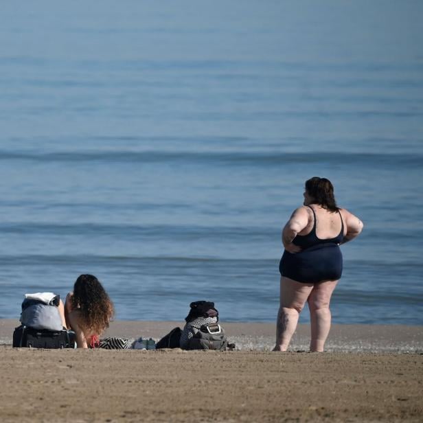 Zwei Frauen mit Taschen am Strand, eine sitzt im Sand, die andere steht im Badeanzug und blickt aufs Meer.