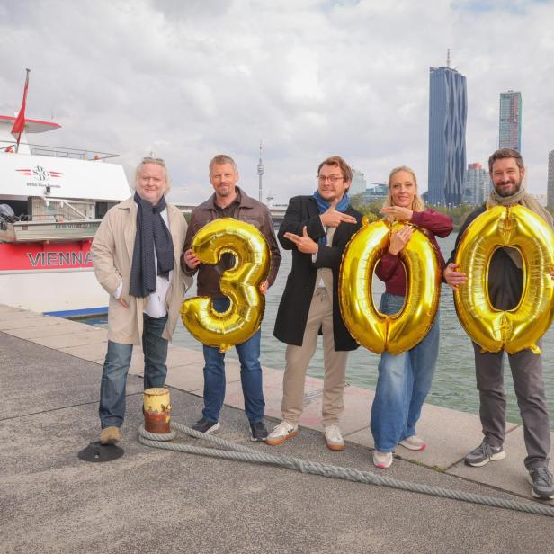 Fünf Personen stehen am Ufer mit goldenen Luftballons in Form der Zahl 300, im Hintergrund ein Schiff und moderne Hochhäuser.