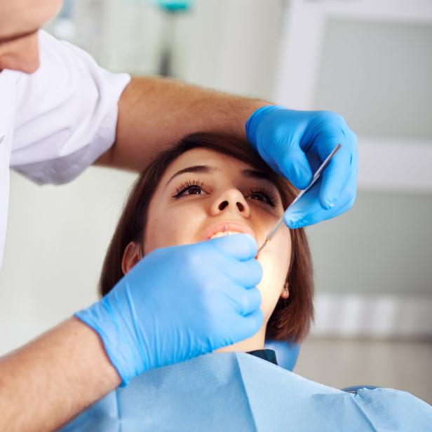 Eine Frau sitzt auf einem Behandlungsstuhl, während ein Zahnarzt mit blauen Handschuhen ihre Zähne untersucht.