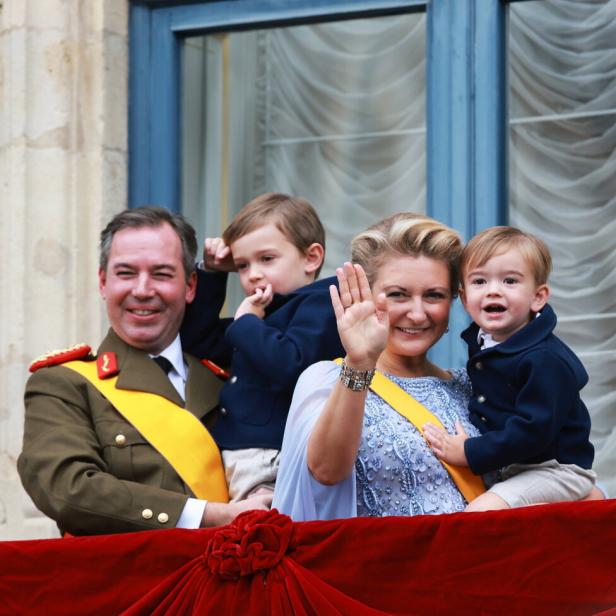 Großherzog Guillaume in Uniform und seine Frau winken von einem Balkon mit roter Balustrade.