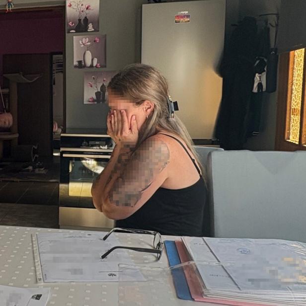 Eine Frau mit Tätowierung sitzt verzweifelt an einem Tisch mit Dokumenten.