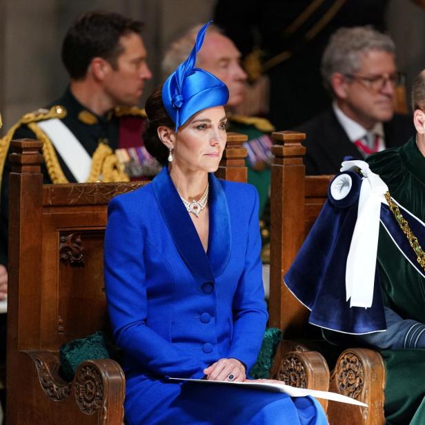 Kate, in blauem Kostüm und Hut, sitzt neben William bei einer Veranstaltung.
