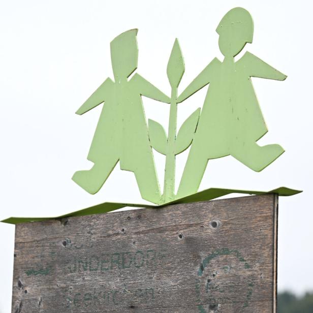 Zwei stilisierte grüne Kinderfiguren halten eine Pflanze auf einem verwitterten Holzschild.