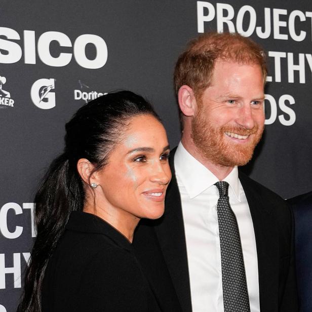 Meghan und Prinz Harry lächeln auf einer Veranstaltung vor einem dunklen Hintergrund.