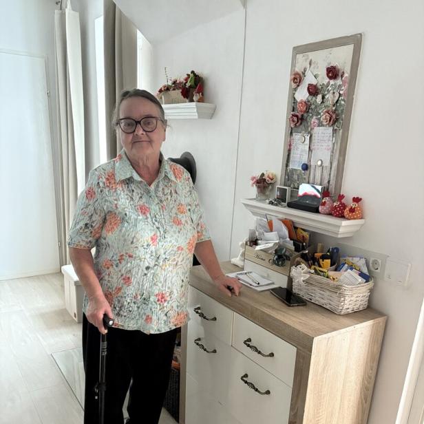 Eine ältere Frau mit Brille und Gehstock steht neben einer Kommode in einem hellen Raum.