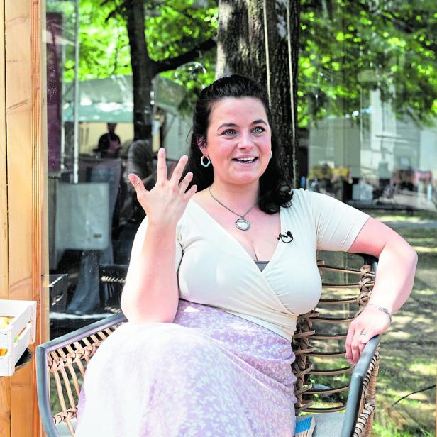 Eine Frau sitzt lächelnd auf einem Stuhl im Freien, gestikuliert mit der Hand und trägt ein Mikrofon am Oberteil.