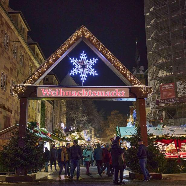 Eingang zum Weihnachtsmarkt mit beleuchtetem Schneeflockensymbol und Schriftzug 'Weihnachtsmarkt', Menschen und Stände bei Nacht.