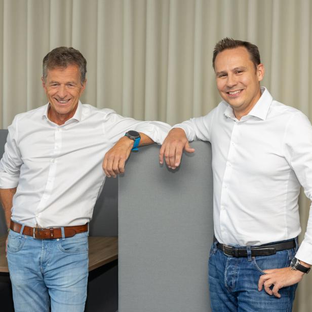 Zwei Männer in weißen Hemden und Jeans stehen lächelnd nebeneinander an einer grauen Trennwand.