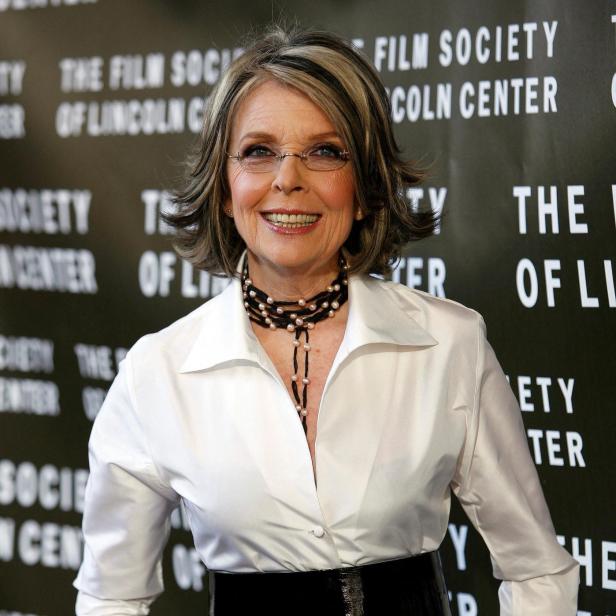 Diane Keaton lächelt in weißer Bluse, schwarzem Gürtel und Perlenkette vor einem schwarzen Hintergrund.