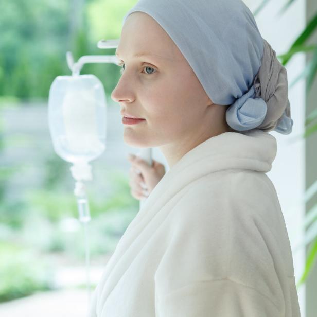 Eine Frau mit Kopftuch und Bademantel steht mit Infusion am Fenster und blickt nach draußen.