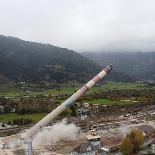 Ein hoher Schornstein stürzt kontrolliert um und wirbelt dabei Staub auf, im Hintergrund grüne Felder und Berge.