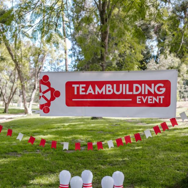 Fünf weiße Bowlingkegel mit roten Ringen auf Gras, darüber ein Banner mit rotem Schriftzug 'TEAMBUILDING EVENT' und roter Grafik, im Hintergrund Bäume.