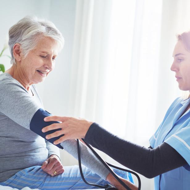 Ältere Frau sitzt, während eine Pflegekraft in blauer Arbeitskleidung ihren Blutdruck misst.