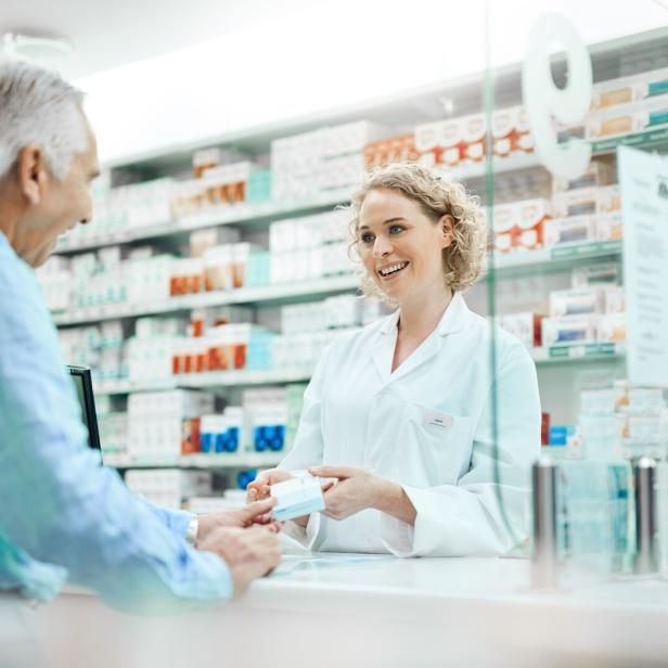 Apothekerin in weißem Kittel übergibt einem Kunden ein Medikament in einer Apotheke mit Regalen voller Arzneimittel im Hintergrund.