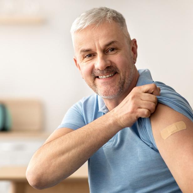 Mann in blauem Poloshirt zeigt Pflaster auf Oberarm, sitzt in heller Küche mit unscharfem Hintergrund.