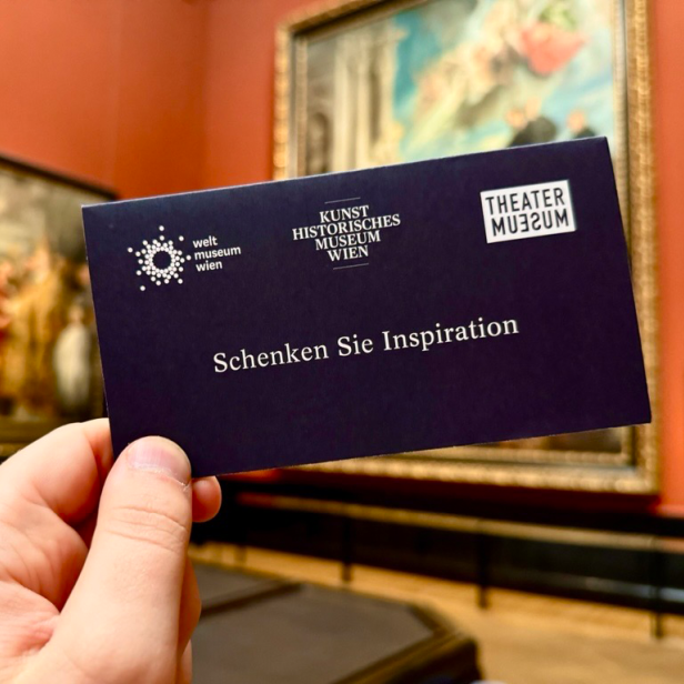 Hand hält dunkelblauen Gutschein mit den Logos von Weltmuseum Wien, Kunsthistorisches Museum Wien und Theatermuseum sowie dem Text 'Schenken Sie Inspiration' vor Gemälden in Museumssaal.